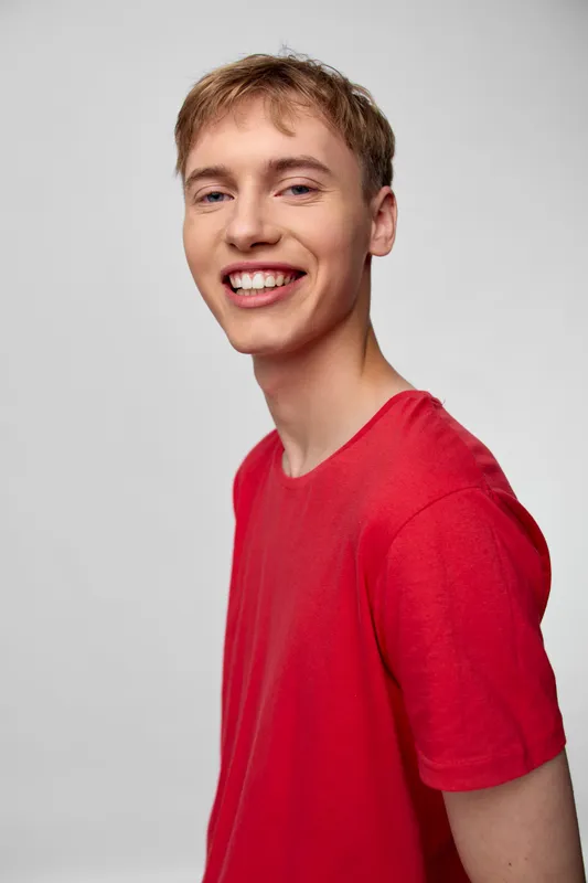 Smiling young man in a red shirt stands in a studio setting, conveying happiness and confidence, casual fashion style, bright lighting, approachable energy, and a positive mood for lifestyle use