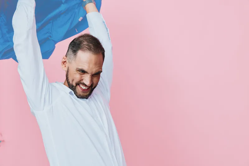 A business professional man stands against a vibrant isolated pink background, displaying confidence, optimism, and energy as he lifts a jacket overhead in celebration.