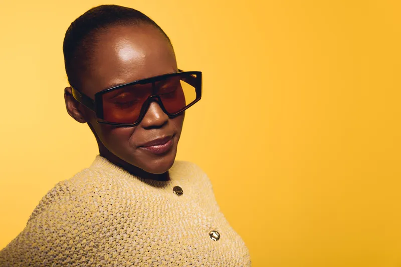 Portrait of confident black African woman with dark skin wearing large sunglasses and beige sweater against yellow background. Concept of fashion, style, and modern elegance.