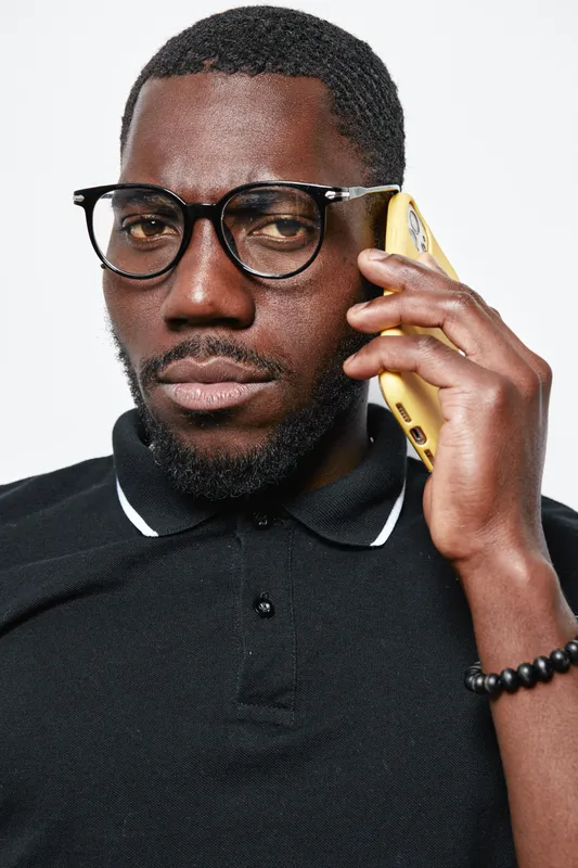 African American man with dark skin wearing glasses and black polo shirt talking on yellow smartphone. Closeup portrait of confident male with beard and bracelet on white background.