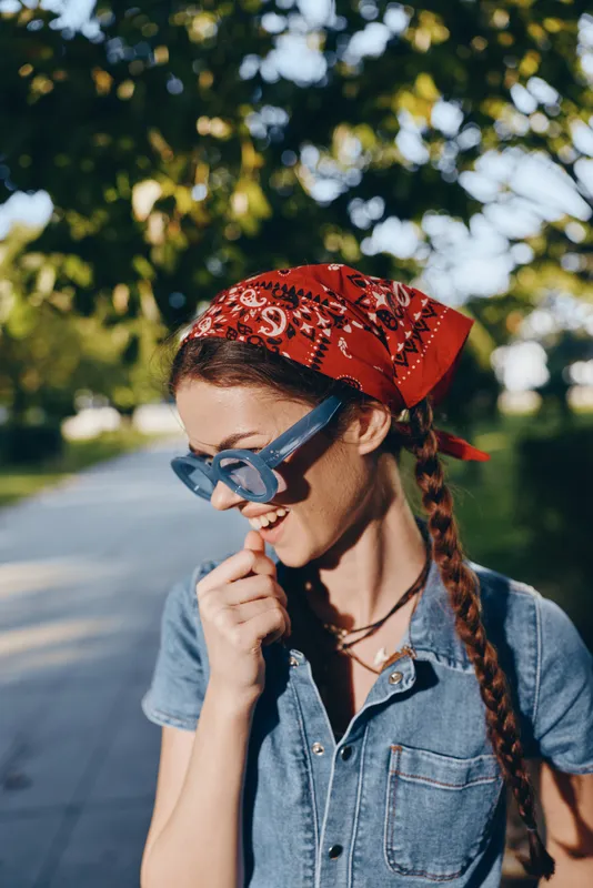 Lifestyle woman in modern boho-western denim with red bandana and blue sunglasses enjoys outdoor street style in warm film color tones, smiling with natural sunlight.