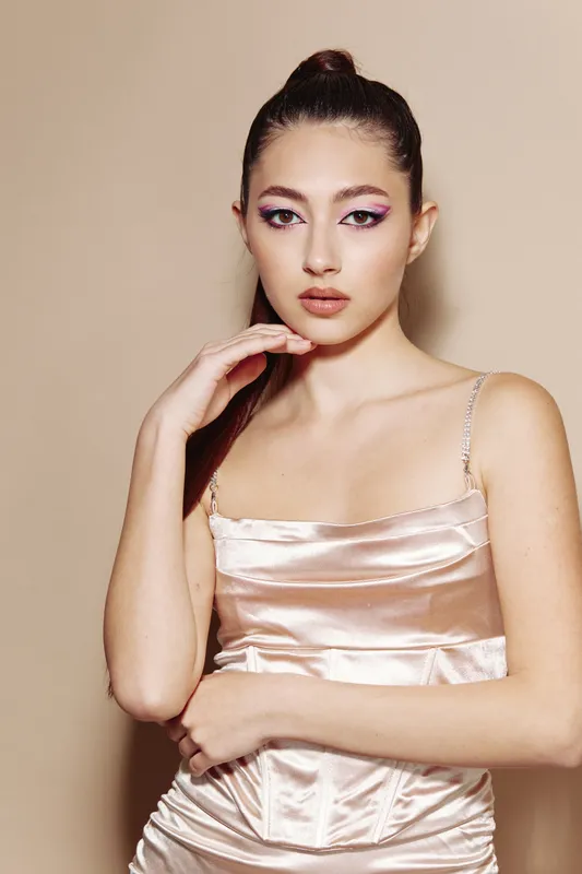 Portrait of young fashion model in satin dress with bold eye makeup and sleek hair. Luxurious styling and artistic pose against neutral background convey creative beauty concept.