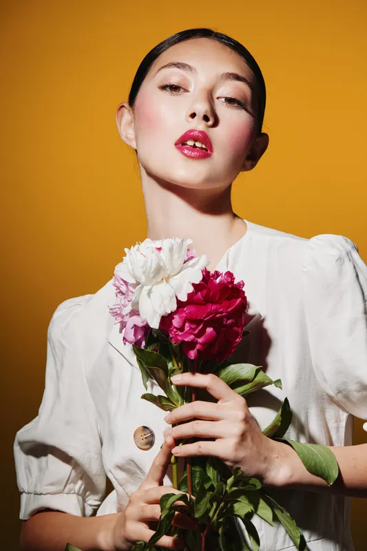 Fashion model with glossy makeup poses holding red and white flowers. Styled with sleek hair and soft lighting on warm orange background. Creative beauty and fashion concept.