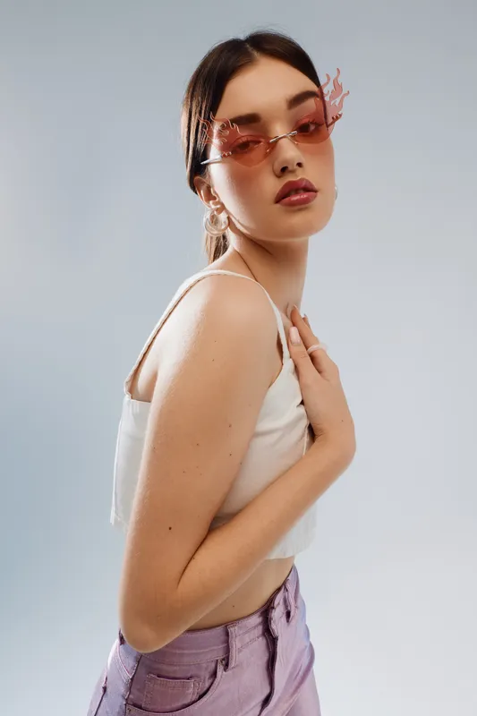 Young woman in pink sunglasses and purple pants poses confidently in a white top against a light background. Concept of beauty fashion and modern style.