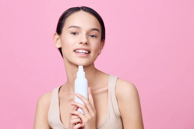 Young woman holding a beauty product with a smile against a soft pink background, showcasing skincare and self care concepts in an elegant style