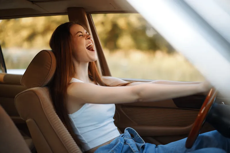 Young woman joyfully driving a car, expressing freedom and happiness on a sunny day with a blurred nature background, wearing a white tank top and blue jeans.