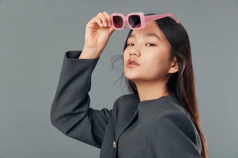 Woman with long hair wearing black jacket holding pink sunglasses near face posing with confident expression on gray background in studio portrait for fashion and style.