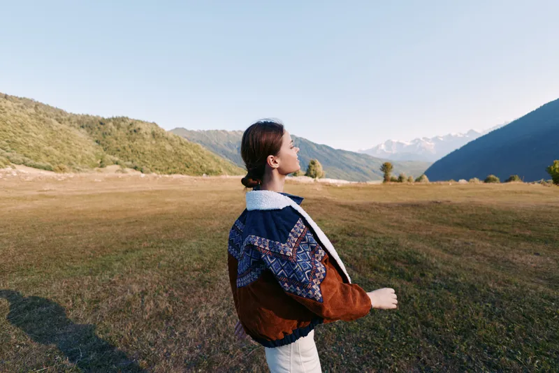 Woman portrait in jacket standing in a wide field landscape, looking toward distant mountains. Nature outdoors at golden hour with valley scenery, serene peaceful travel mood.