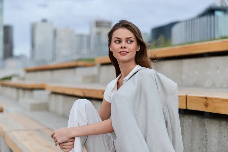 Young woman sitting outdoors in casual white outfit with light jacket, modern urban background, natural smile, relaxed and confident atmosphere