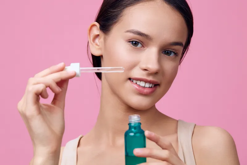 Young woman holding a dropper bottle of serum for skincare, smiling against a pink background, showcasing healthy glow and beauty routine