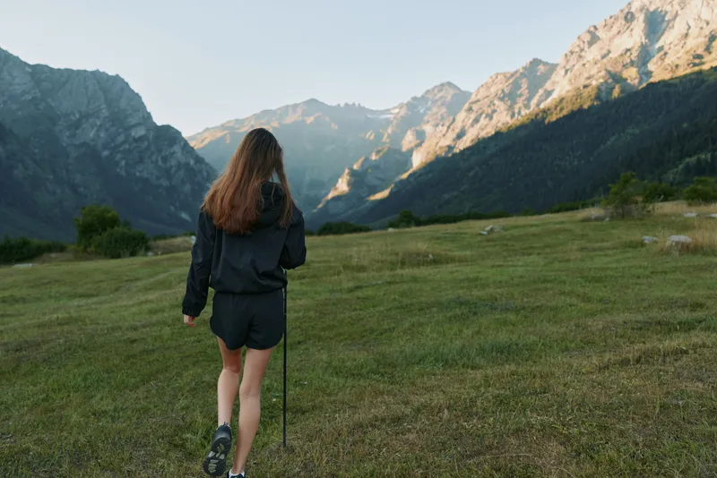 A lone female hiker walks through a grassy valley toward rugged mountains. She wears a dark jacket and shorts, uses trekking poles, exploring a peaceful alpine landscape.