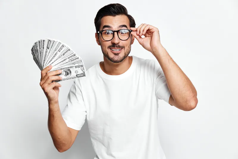 Happy smiling man wearing glasses holding cash money in hand, celebrating financial success, isolated on plain white background in studio portrait. Lifestyle concept.