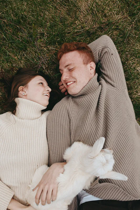 Smiling couple lying on the grass in knit sweaters, a playful goat kid nestled between them. Cozy lifestyle moment, rustic setting, warm sun, and gentle affection conveying care and connection.