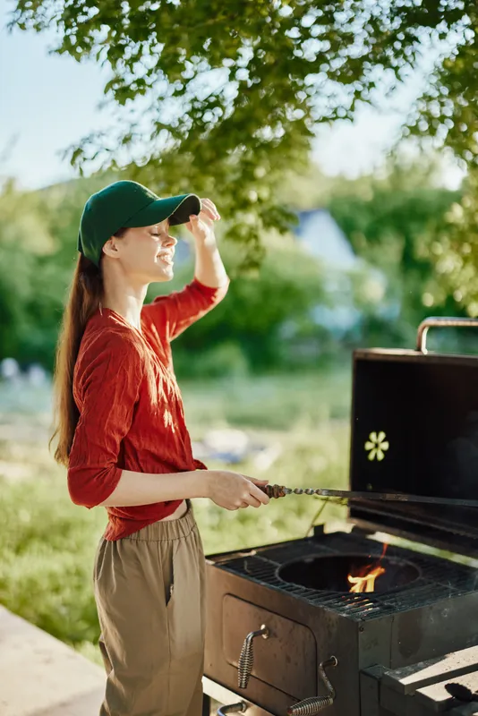 Young woman grilling outdoors in a green cap, smiling joyfully Natural background and warm colors create a summer vibe, ideal for cooking and outdoor activities