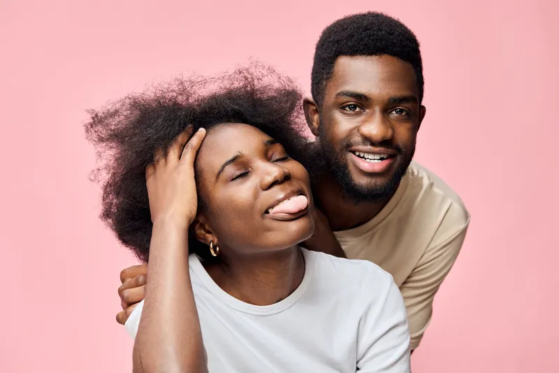 Happy young couple expressing joy with playful emotions on a pink background, showcasing a casual style in summer clothing and natural hairstyles