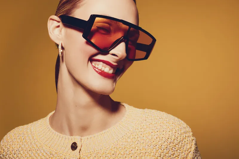 Smiling young woman with short hair wearing oversized orange sunglasses and a knitted beige sweater. Studio portrait on warm brown background. Concept of fashion, style, and happiness.