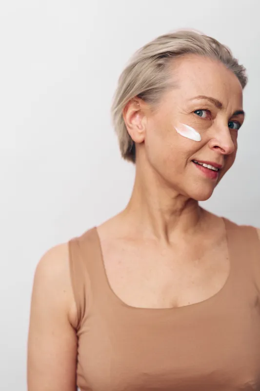 Mature woman with short grey hair applies facial cream on cheek, smiling gently against neutral background. Concept of skincare, aging, wellness, and natural beauty care routine.