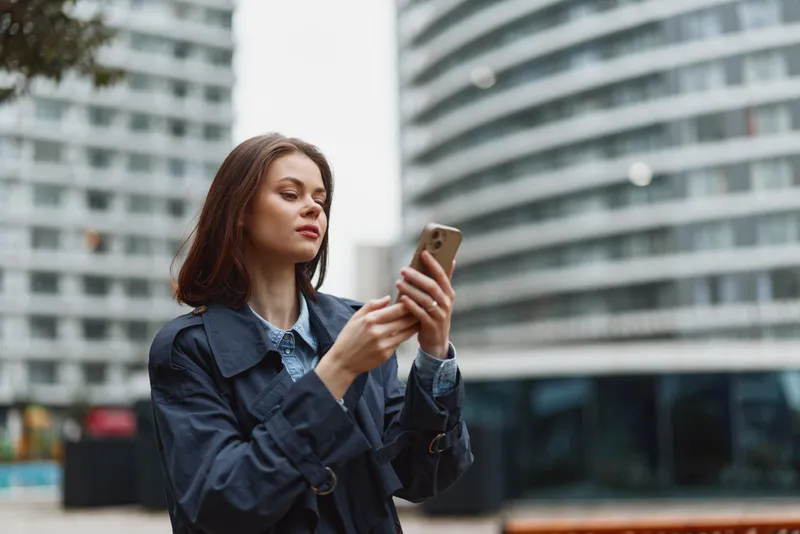 Confident woman outdoors in an urban setting, checking a smartphone, stylish jacket, soft daylight, modern architecture in the background, candid portrait with calm mood