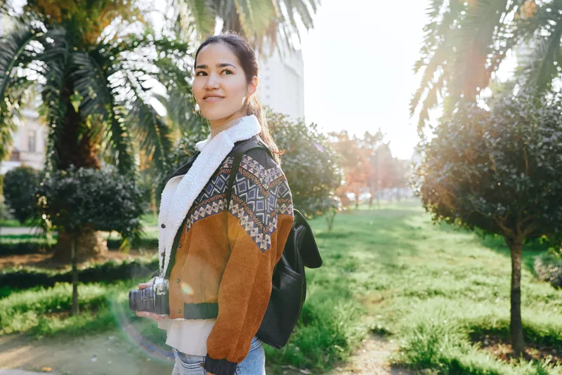 Woman with camera in a sunny park, wearing a color cardigan and scarf, backpack on shoulder, exploring lush greenery and palms, capturing candid moments outdoors today
