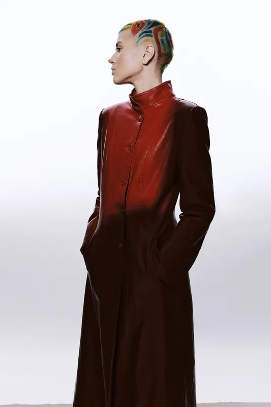 Woman with colorful braided hair wearing a long red coat standing in profile on a white background, showcasing modern fashion and bold style expression.