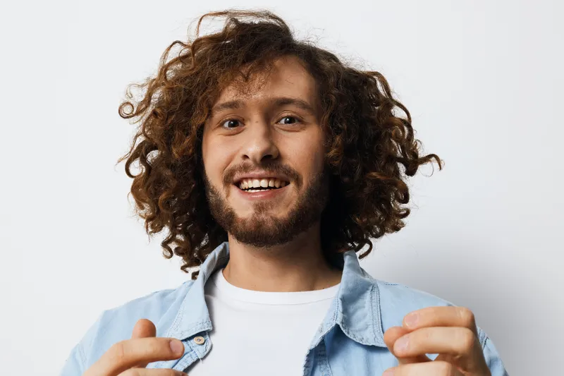 Smiling man with curly hair in denim jacket expressing joy and positivity against a light background, ideal for promoting happiness and connection