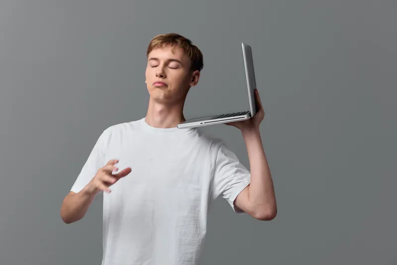 Young man in a white t shirt balances a laptop on his shoulder, mid gesture, illustrating multitasking, casual work, and tech use in a modern office or home workspace