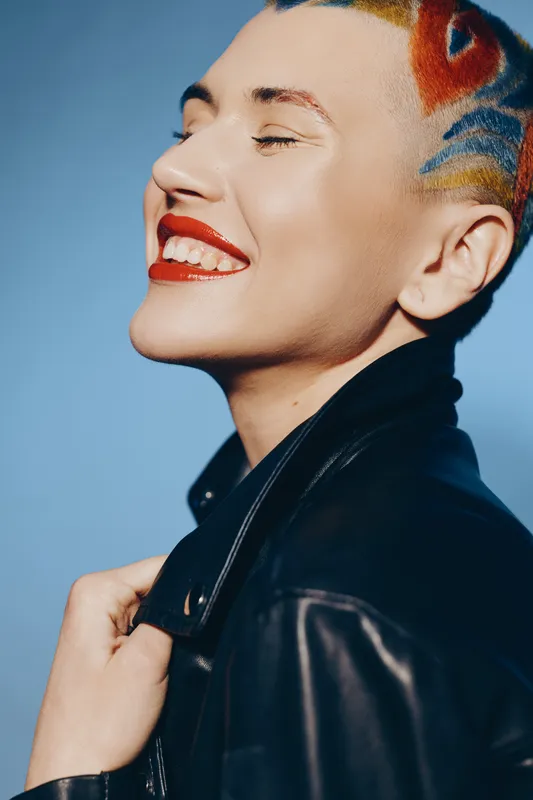 Smiling woman with short colorful hair wearing a black leather jacket and vibrant red lipstick, expressing joy against a soft blue background in natural light.