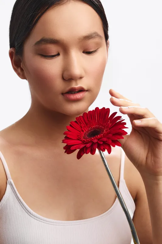 asian woman studies a vivid red flower, close up portrait with soft lighting that highlights her calm features, creating a serene mood and graceful presence.