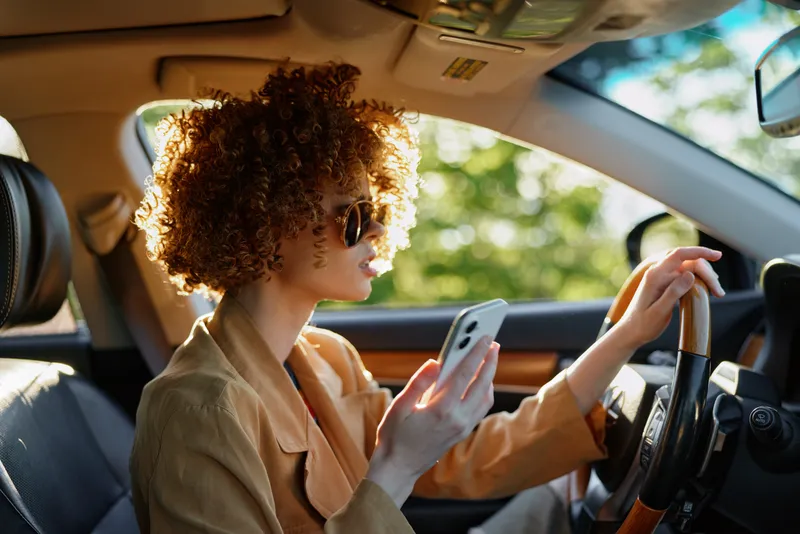 Casual woman driving a car, talking on a phone, wearing sunglasses, sunlit interior with warm retro tones, relaxed mood, leafy view through the window