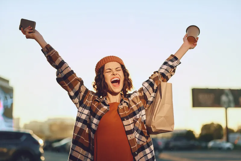 Woman joyfully raising hands with coffee and shopping bag in urban morning, candid lifestyle portrait radiating authenticity, golden hour glow, mindful living and emotional storytelling.
