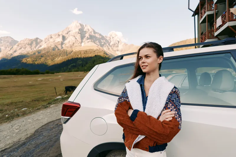woman car mountains travel roadtrip outdoor portrait nature. Young woman leans against white SUV on rural roadside with scenic mountain backdrop, casual sweater and relaxed adventurous expression.