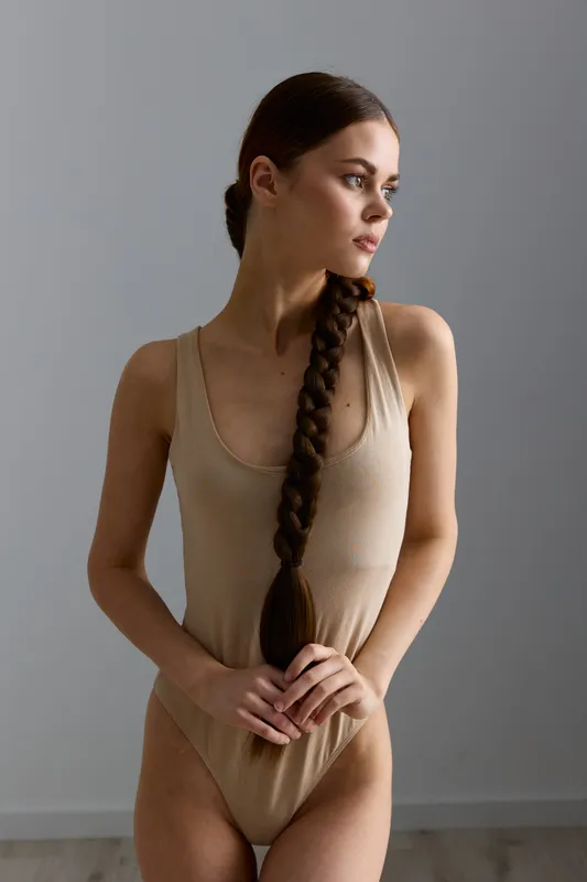Young woman with long braided hair in beige bodysuit poses thoughtfully against neutral background, showcasing natural beauty, minimalistic style, and calm mood in soft lighting.