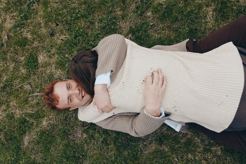 Cheerful couple outdoors, lying on grass, embracing and smiling, sunlit scene, casual outfits, soft breeze, natural colors, relaxed mood, candid moment, affectionate connection