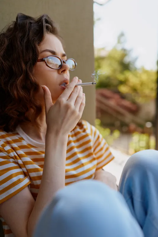 Casual woman with glasses sits outdoors on a sunny day, wearing striped tee and jeans, smoking a cigarette with a pensive relaxed mood, natural light, candid moment