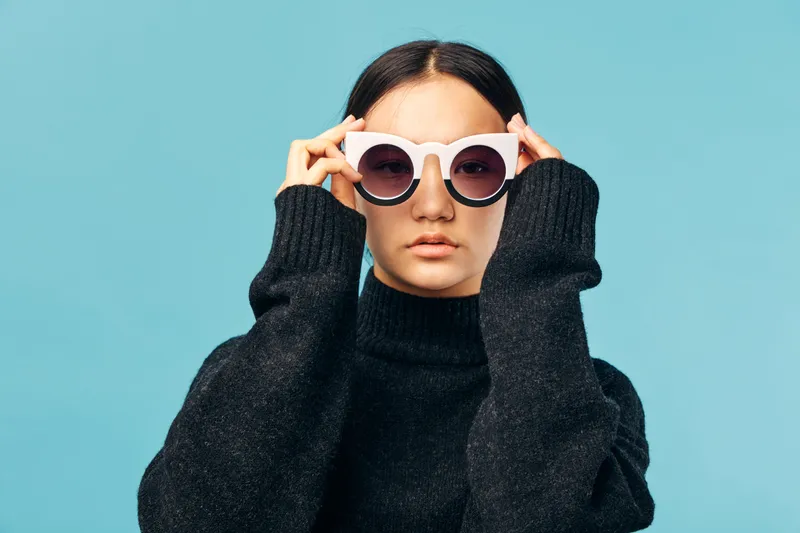 Woman with round sunglasses and black sweater posing on blue background looking forward with serious expression and long hair tied back in studio photo.