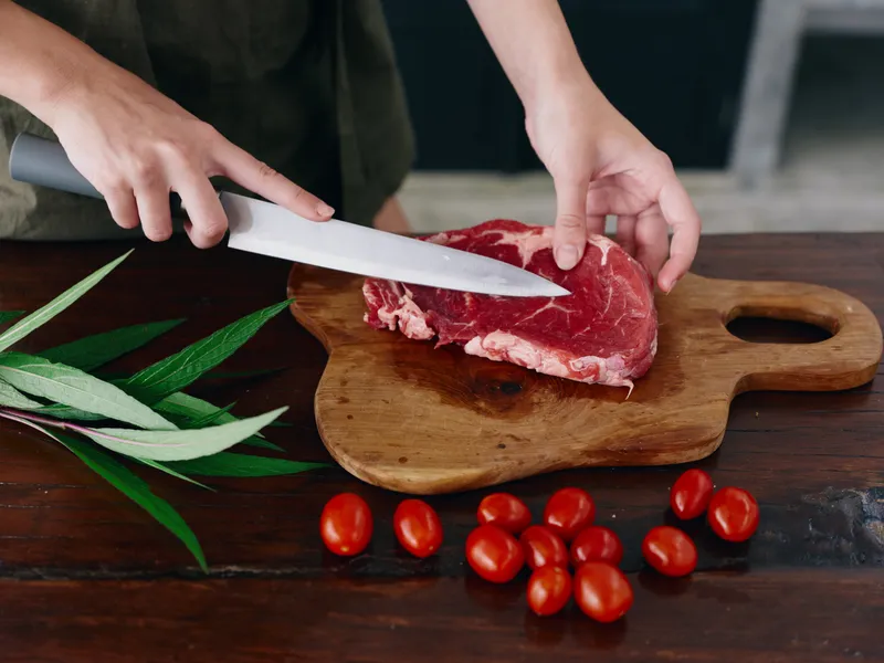 Женщина с ножом в руке нарезает свежее мясо стейка для запекания на кухне, на столе соль, перец и другие специи, красные помидоры черри и зелень, готовит ужин