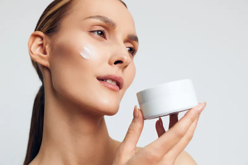 Woman applying cream on her face, holding a white jar, close up portrait, skincare and beauty concept, soft studio light on neutral background, selfcare wellness, daily care