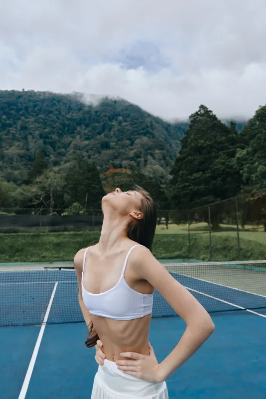 Young female athlete enjoying fresh air on a tennis court, wearing a white sports top and skirt, showcasing confidence and relaxation in nature