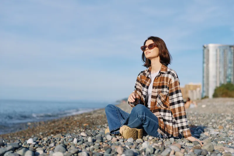 Woman on a pebble beach wearing a plaid shirt and jeans, enjoying sunny seaside moments with sunglasses, relaxed pose near the water and distant cityscape.