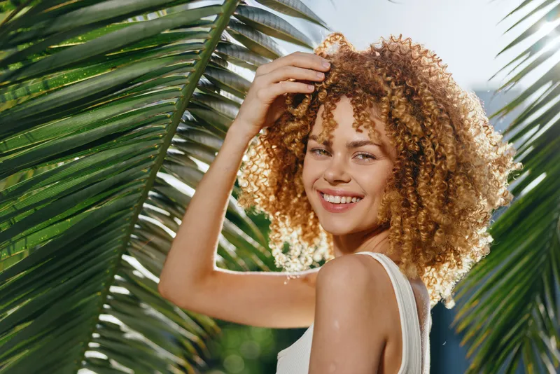 smiling woman with curly hair poses joyfully in sunlight amidst tropical palm leaves, wearing a white tank top and natural makeup, bright summer mood