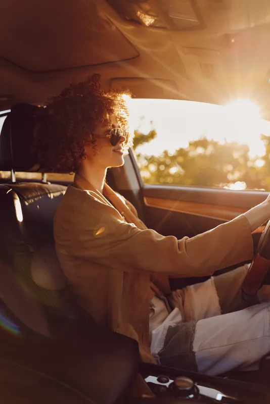 Young woman with curly hair drives a car at sunset, wearing sunglasses and a cozy knit, conveying a retro road trip mood and warm, hopeful energy.