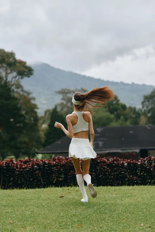 Young female athlete running in a white athletic outfit on a green field, showcasing energy and joy, with trees and mountains in the background