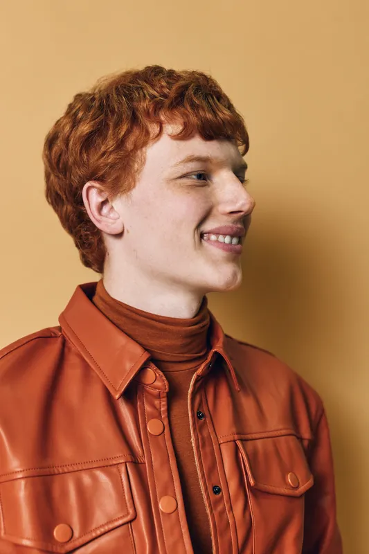 Young man with red hair and freckles smiling in brown leather jacket and turtleneck sweater against beige background in studio portrait.
