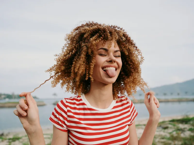 Playful portrait of a smiling woman with curly hair sticking out her tongue, wearing a red and white striped shirt, outdoors by a calm lake with distant mountains.