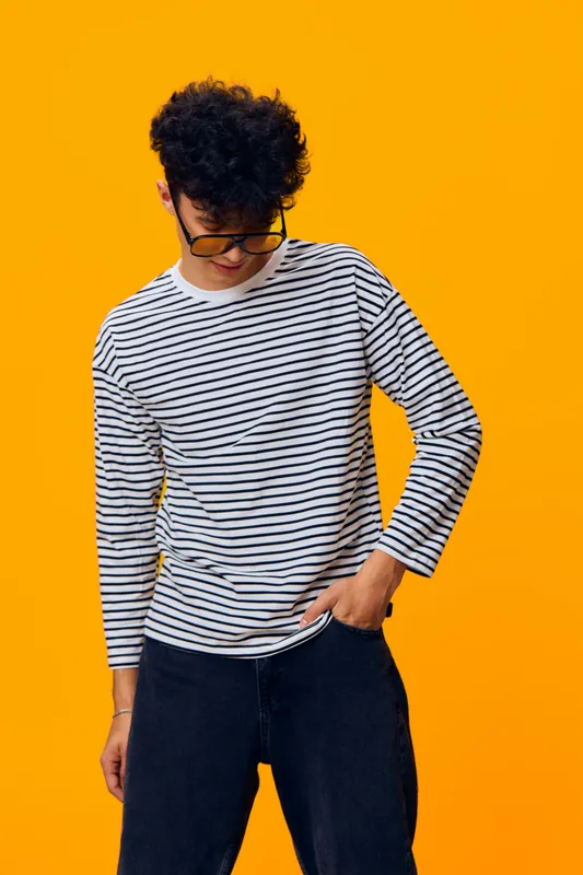 young man wearing striped shirt and glasses stands casually against bright orange background, looking down, with curly hair and relaxed pose