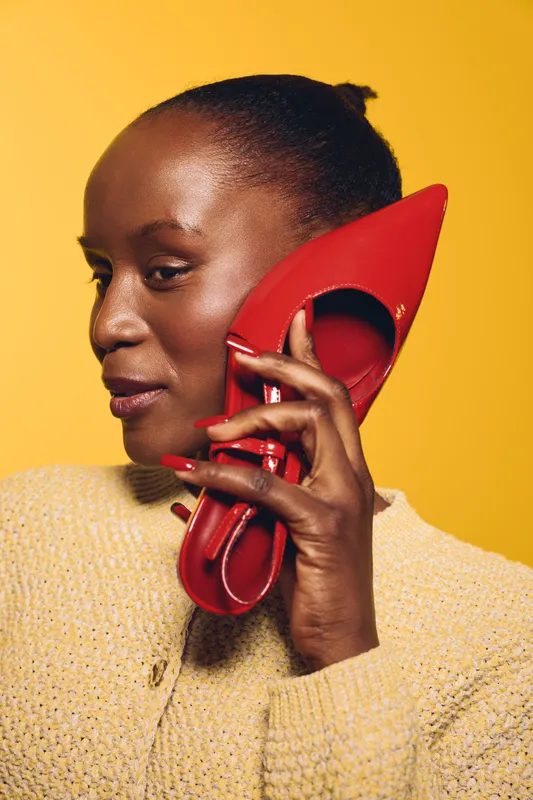Portrait of a dark skinned African woman holding a red high heel shoe to her ear like a phone. Yellow background and beige sweater create a bold, creative fashion concept with playful mood.