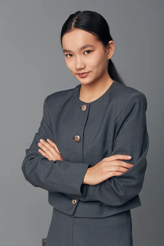 Portrait of confident young woman with folded arms wearing gray formal jacket standing against plain background looking at camera with slight smile in studio setting.