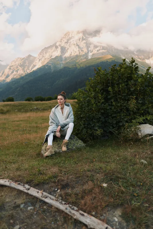 Young woman in casual clothes sitting on rock near green bush with mountain landscape in background. Concept of outdoor lifestyle, relaxation, and nature escape.