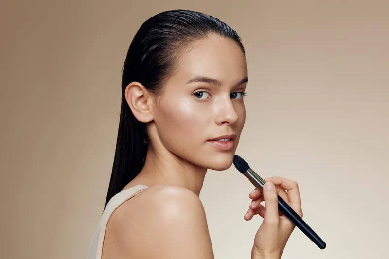 Portrait of a young model applying makeup with a brush against a soft beige background, showcasing natural beauty and confidence