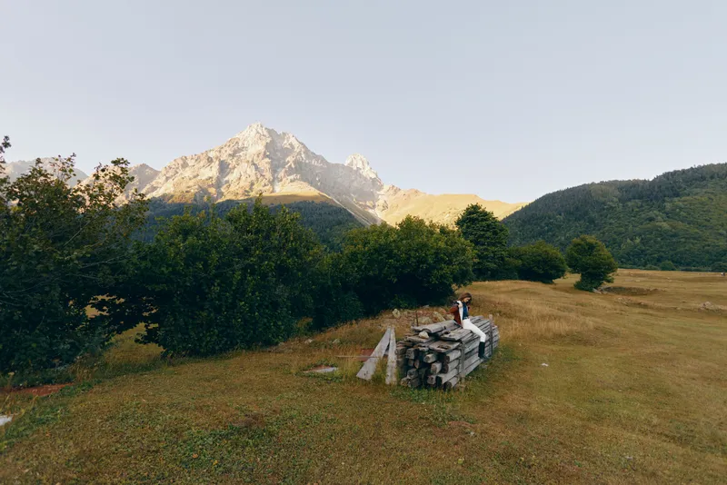 Mountain meadow with woodpile and person sitting near trees, landscape view of nature and hiking trail at sunrise, peaceful alpine field and rustic outdoor scenery.